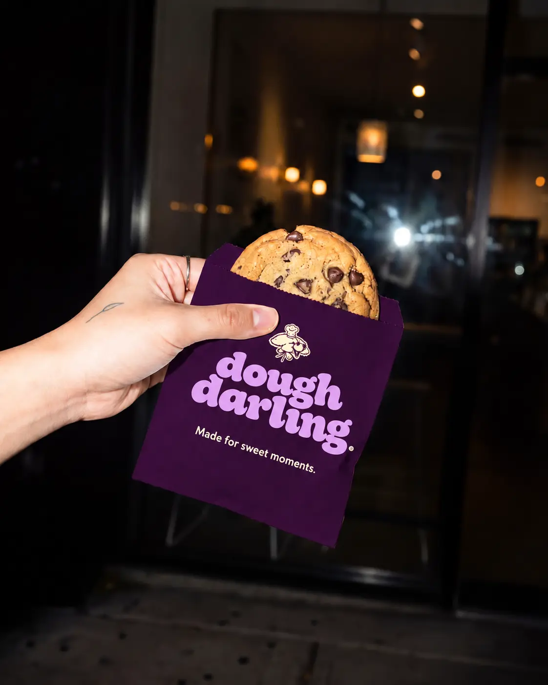 A hand holding a warm chocolate chip cookie in a deep plum Dough Darling sleeve