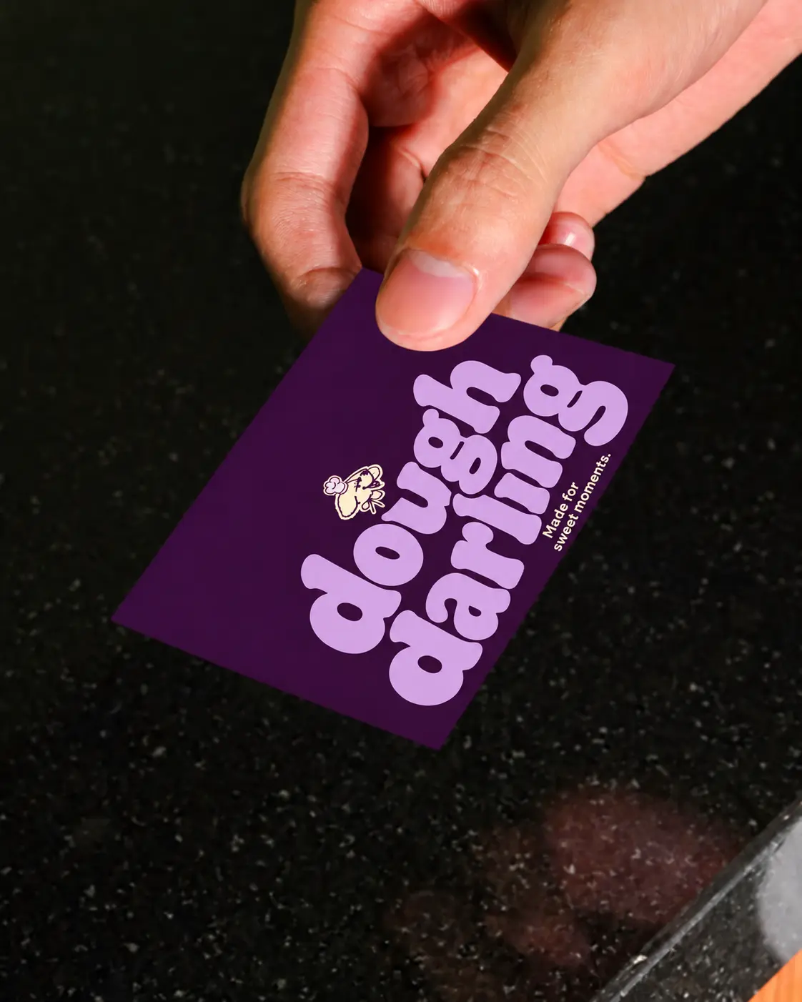 A hand holding a Dough Darling business card with lilac retro lettering